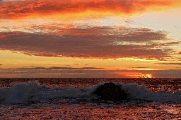 Painted sky of a brilliant sunset over wave crashing past a sea rock seen in silhouette